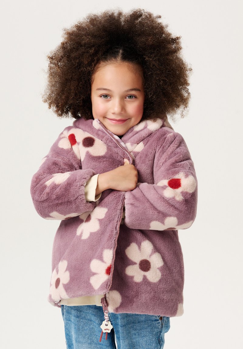 Paarse fleecejack met grote witte en rode bloemmotieven, ritssluiting aan de voorkant en elastische manchetten, gecombineerd met een lichtgekleurde top en denim.