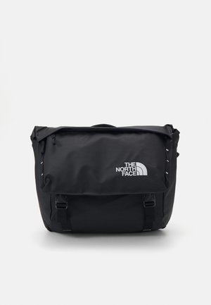 Bolso mensajero negro hecho de un tejido duradero, con una solapa doblable, correas ajustables y un logo blanco en la parte delantera.