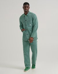 Green corduroy jacket with front pockets, button closure, paired with matching pants. Person wears green shoes, stands against a neutral background.