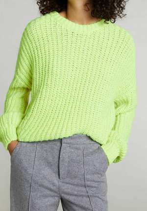 Persona che indossa un maglione spesso verde lime a maglia con polsini a coste e pantaloni grigi a vita alta, mani nelle tasche, capelli ricci visibili.