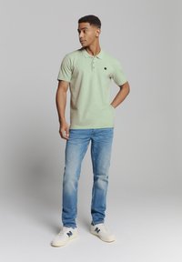 Lichtgroen polo shirt met een tweeknoopsluiting, gedragen met blauwe jeans en witte sneakers met een blauw logo. Eenvoudige, casual stijl.