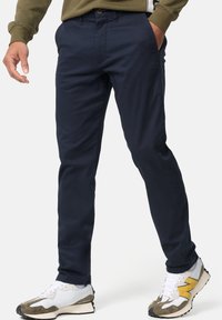 Chino bleu marine en coton, design ajusté, devant plat avec poches, doté d'un bouton marron et d'une texture lisse et mate.