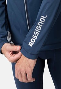 Marinblå prestationsjacka med grå detaljer och vit "ROSSIGNOL"-logotyp på ärmen. Slät, stretchig tyg med framsidan dragkedja och tajt passform.