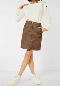 Jupe en denim marron avec poches avant, fermeture par boutons et détails de couture. Associée à un haut à manches longues blanc et des baskets blanches.