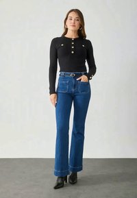 Črn rebrast dolg rokav vrh z okroglim izrezom in gumbi, kombiniran z modrimi razširjenimi jeans hlačami in črnimi visokimi gležnarji.