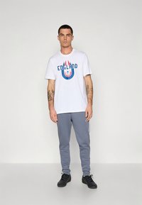 Biele bavlnené tričko s modro-červenou grafikou "ENGLAND", doplnené sivými teplákmi s bielymi bočnými pruhmi. Čierne športové topánky.