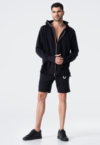 Czarna bluza z kapturem zapinana na zamek, z wysokim kołnierzem, warstwowym dołem i długimi rękawami; w połączeniu z czarnymi szortami z okrągłym logo. Czarne sneakersy.