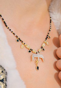 Collier avec des perles noires, des accents dorés et un pendentif en argent en forme d'oiseau avec une gemme noire en forme de goutte. Designs et textures complexes mis en valeur.