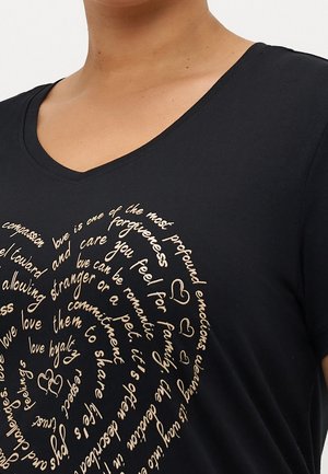 Person iført en sort V-hals skjorte med beige spiraltekst om kærlighed, inklusive hjerter og ord som tilgivelse og loyalitet.