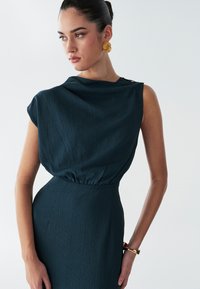 Robe bleu sarcelle en tissu texturé ; présente un col drapé, de légères manches bouffantes et une taille cintrée. Boucles d'oreilles dorées accrocheuses et bracelet.