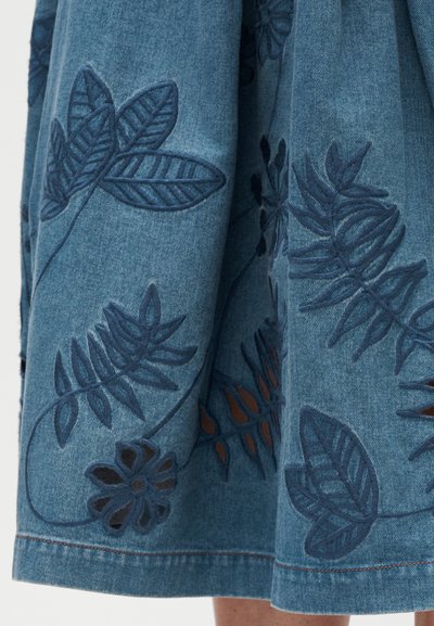 Jupe en denim bleu avec des motifs floraux et de feuilles brodés en bleu foncé, présentant des détails découpés et des coutures contrastantes le long de l'ourlet.