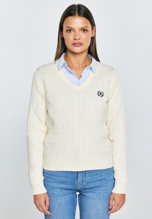 Krémový svetr s výstřihem do V a copánkovým vzorem s žebrovaným lemem a manžetami, s vyšívaným logem na levé straně hrudi. Nosit přes světle modrou košili s límečkem.