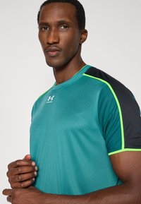 Türkizkék sportpóló fekete ujjakkal és zöld díszítéssel. Simai anyagból készült, kerek nyakkal és az Under Armour logóval az elején.