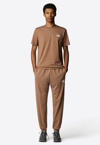 Bruine katoenen t-shirt en bijpassende joggers met elastische boorden, voorzien van een klein wit logo op de borst en bovenbeen. Zwart en grijs gemêleerde sneakers.