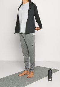Sudadera con cremallera negra, camiseta gris y pantalones de chándal grises. Tejido ligero, corte relajado, con el logo de Nike en los pantalones. Botella de agua al lado.