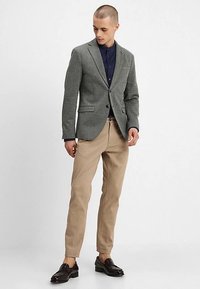 Szary teksturowany blezer z wciętymi klapami, noszony na ciemnoniebieskiej koszuli, w zestawie z beżowymi spodniami i czarnymi butami. Czyste, dopasowane krawiectwo.