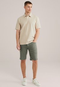 Svetlo bež polo majica s kratkimi rokavi in teksturiranim vzorcem, kombinirana z olivno zelenimi kratkimi hlačami. Model nosi bele superge.