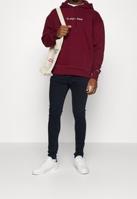 Felpa bordeaux con testo bianco "Tommy Jeans", abbinata a jeans skinny scuri e sneaker bianche, mentre porta una borsa a tracolla beige.