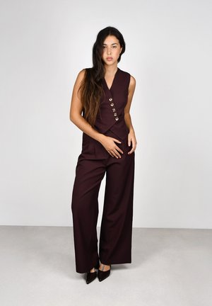 Joven con el cabello largo, vistiendo un chaleco burdeos sin mangas con botones y pantalones anchos a juego, de pie contra un fondo liso.