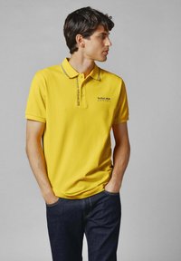 Geltona polo marškinėliai su juodais akcentais, su logotipu kairiajame krūtinės šone. Trumpomis rankovėmis, standartiniu apykaklę, derinami su tamsiomis džinsinėmis kelnėmis.