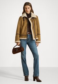 Giacca marrone foderata in shearling con zip frontale, di colore beige chiaro e con dettagli in pelle, abbinata a jeans blu e stivali con tacco marroni.