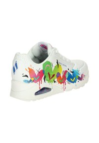 Baskets blanches Skechers avec des cœurs colorés et des gouttes style graffiti sur le côté, coussin d’air visible dans la semelle et logo bleu sur le talon.