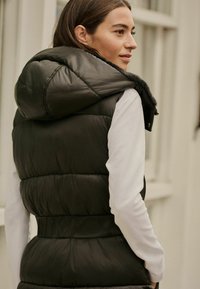Vesta neagră captusită cu glugă, având un design cu quilt și talie strânsă. Purtați-o peste un tricou alb cu mânecă lungă.