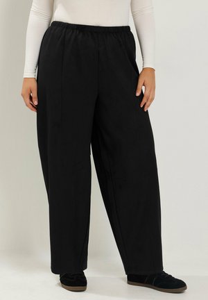 Persoon draagt losse zwarte broek met elastische tailleband, witte lange mouw top en zwarte schoenen, staand tegen een effen achtergrond.