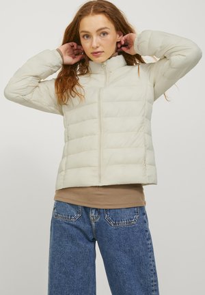 Giubbotto puffer off-white con colletto alto, caratterizzato da pannelli trapuntati orizzontali, una zip frontale e polsini elasticizzati. Indossato sopra un top marrone.