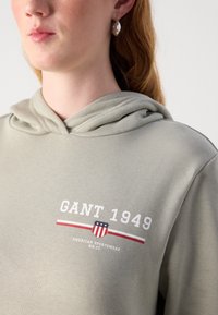 Sudadera verde claro con una textura suave, que presenta un estampado gráfico en blanco y rojo. Incluye una capucha con cordón y sutiles detalles de costura.