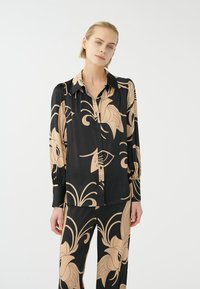 Zwarte zijden blouse met grote beige bloemenpatronen, knoopsluiting aan de voorkant, puntige kraag en lange mouwen. Gepaard met bijpassende broek.