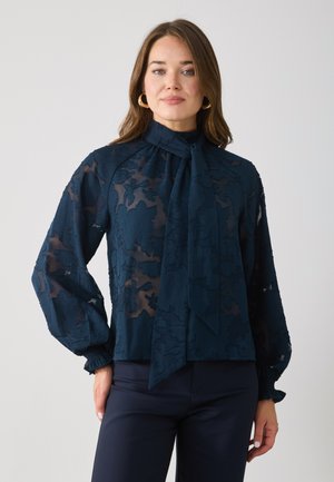 Mørkeblå gjennomsiktig bluse med blomstermønster, høy hals og knytedetalj; har ballongsleeves og innsamlede mansjetter.