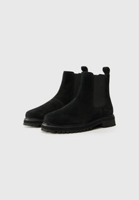 Botines de ante negro con paneles laterales elásticos y lengüetas en la parte superior. Suelas de goma gruesa con banda de rodadura distintiva para tracción.