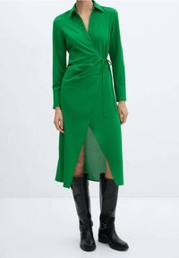 Grünes Wickelkleid mit langen Ärmeln, Kragen, Schlitz vorne und Bindegürtel in der Taille, kombiniert mit kniehohen schwarzen Stiefeln. Glatte Stofftextur.