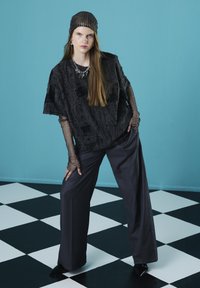 Zwarte, gestructureerde oversized top met lange mouwen van gaas, gecombineerd met grijze wijde broek. Model draagt een zwarte gaas hoofddoek en accessoires.