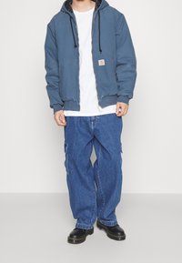 Persoon in een blauwe rits hoodie, witte T-shirt, losse blauwe spijkerbroek en zwarte laarzen, staand tegen een effen lichte achtergrond.