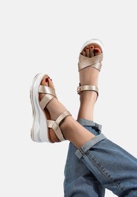 Sandali con plateau a forma di cuneo con una finitura metallica dorata lucida, caratterizzati da cinturini incrociati e una suola bianca. Nel retro si vedono dei risvolti di jeans.