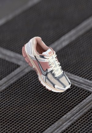 Nike hardloopschoen in beige, roze en grijs met zwarte swoosh, zwevend boven een metalen rooster vloerachtergrond.
