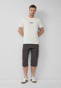 T-shirt blanche à manches courtes avec un logo bleu, assortie à un short cargo gris foncé et des baskets blanches avec des accents beiges. Tenue simple et décontractée.