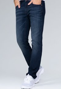 Jeans de mezclilla azul oscuro con corte slim, que presentan un diseño clásico de cinco bolsillos y detalles de costura sutiles. Combinados con zapatillas blancas.