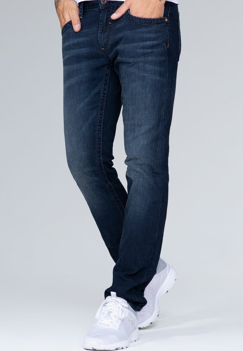 Jeans de mezclilla azul oscuro con corte slim, que presentan un diseño clásico de cinco bolsillos y detalles de costura sutiles. Combinados con zapatillas blancas.