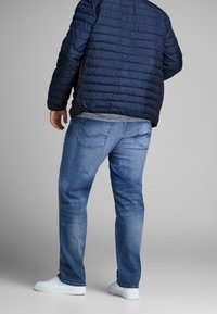 Padded navy jacket with horizontal stitching, light gray shirt underneath, paired with blue denim jeans and white sneakers.