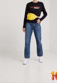 Felpa nera con logo rosso, jeans blu, sneakers bianche e una borsa in vita gialla. La borsa presenta un tessuto con trama e una tracolla regolabile.