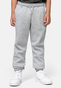 Niño con pantalones de chándal gris claro y cintura con cordón, vistiendo zapatillas blancas, de pie contra un fondo blanco liso.