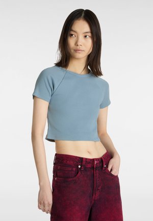 T-shirt cropped di colore blu chiaro realizzato in tessuto a coste, con maniche corte, scollatura rotonda e forma aderente. Abbinato a jeans rossi a vita alta.