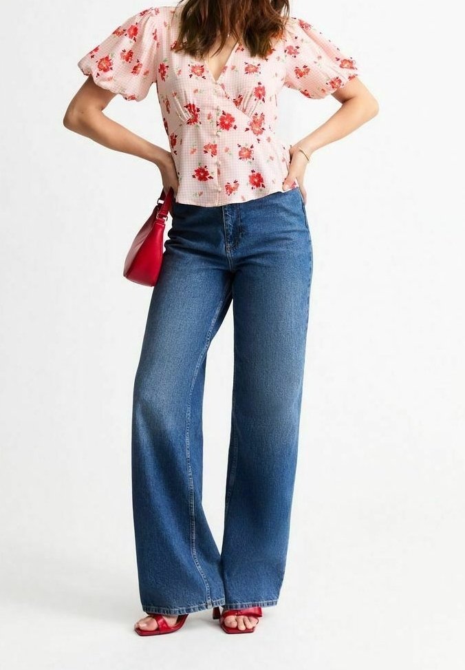 Femme portant un chemisier rose à manches bouffantes à motifs floraux, un jean bleu large taille haute, un sac à main rouge et des talons ouverts rouges, debout les mains sur les hanches.
