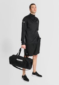 Schwarzes langärmeliges Sportoberteil mit hohem Kragen, schwarze Shorts und schwarze Sneakers. Hält eine schwarze Sporttasche mit dem weißen "HUMMEL"-Logo.