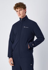 Marineblå glidelåsjakke laget av mykt stoff. Har stående krage, to sidelommer, og en liten logo på brystet og ermet.