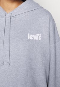 Osoba oblečená v světle šedé mikině Levi's s viditelným logem na hrudi a šňůrkami na kapuce spadlými dopředu.