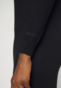 Primo piano di una mano di carnagione scura che indossa un indumento a maniche lunghe nero con il sottile logo "Calvin Klein" ricamato vicino al polso.
